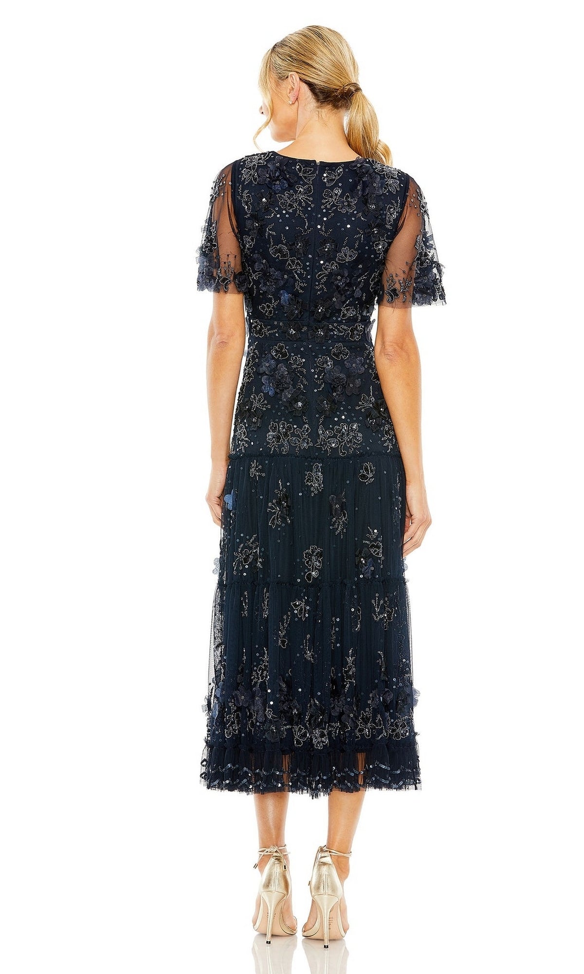 A woman stands with her back to the camera, wearing the Mac Duggal 9207 A-Line Wedding Guest Dress in navy blue with floral embellishments, short sheer sleeves, and metallic heels.